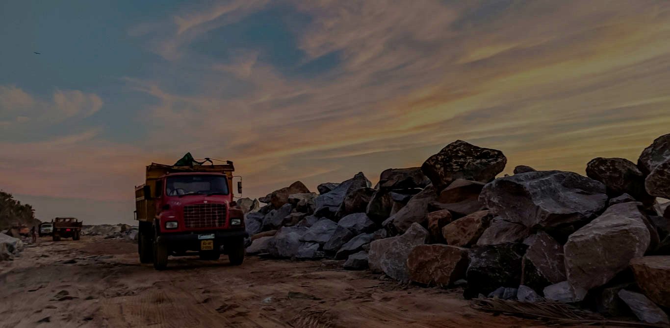 A red tipper truck beside a stockpile of boulders on a site road at dusk